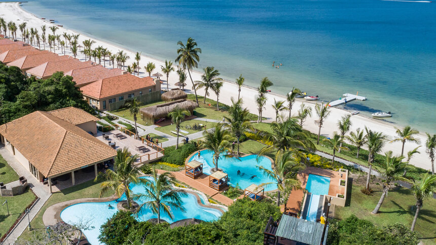 Aerial view of a tropical beachfront resort with swimming pools, palm trees, beachfront villas, and a white sandy beach beside clear blue ocean waters.